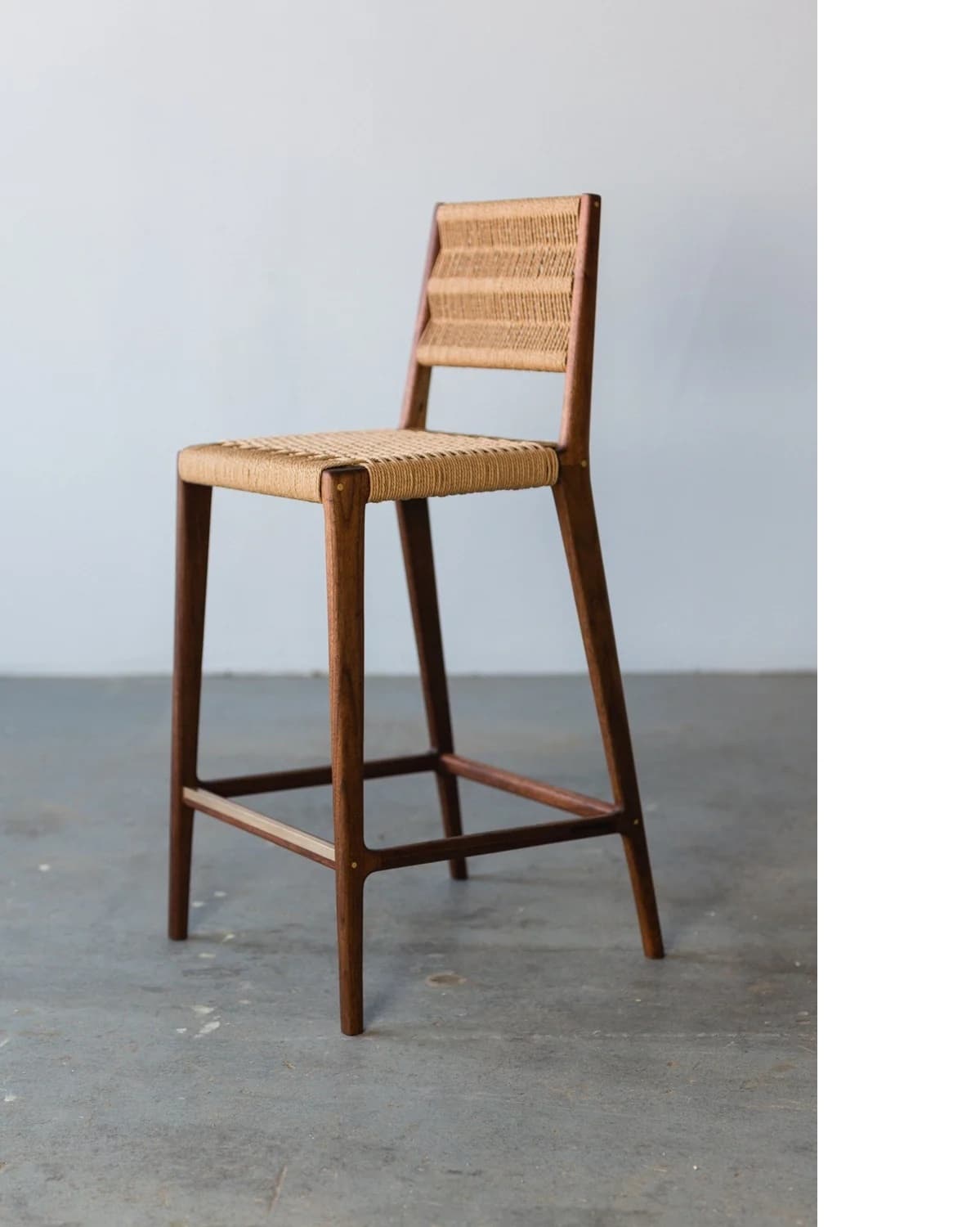A bar stool with a wooden frame and a woven seat and backrest. The stool features four tapered legs connected by a lower stretcher for stability. The seat and backrest are constructed from a natural woven material, creating a textured surface. The wooden frame appears to be dark-stained. — 3D model