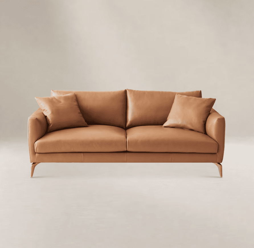 A modern two-seater sofa featuring clean lines and upholstered in smooth tan or cognac-colored leather. It includes two plush seat cushions, two back cushions, and two matching square throw pillows. The sofa is supported by four slender, tapered legs with a polished brass finish. — 3D model