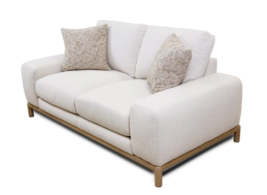 A modern, two-seater sofa featuring a plush, textured cream or off-white fabric upholstery. It has gently rounded track arms and a low back with two removable back cushions. The seating area consists of two separate seat cushions. The sofa is elevated by a minimalist wooden frame that forms the base and extends into four short, cylindrical wooden legs. Two smaller, patterned throw pillows are included. — 3D model