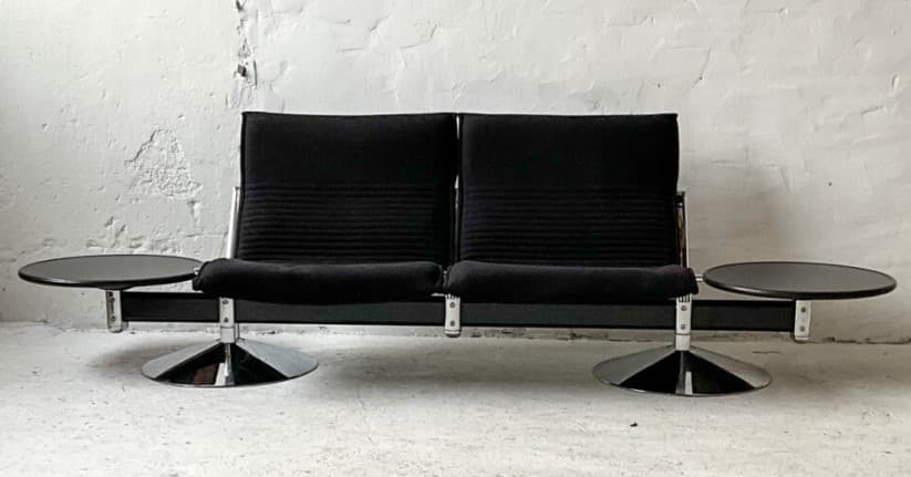 A two-seater sofa with attached side tables. The sofa features black upholstered seats and backrests with a ribbed texture on the backrests. The frame appears to be metal, possibly chrome or stainless steel, with a dark finish on the main horizontal support. Each seat has a cantilevered base with a circular, flared chrome pedestal. Attached to the horizontal frame on either side are circular, dark-colored side tables with a glossy finish, supported by metal brackets. — 3D model