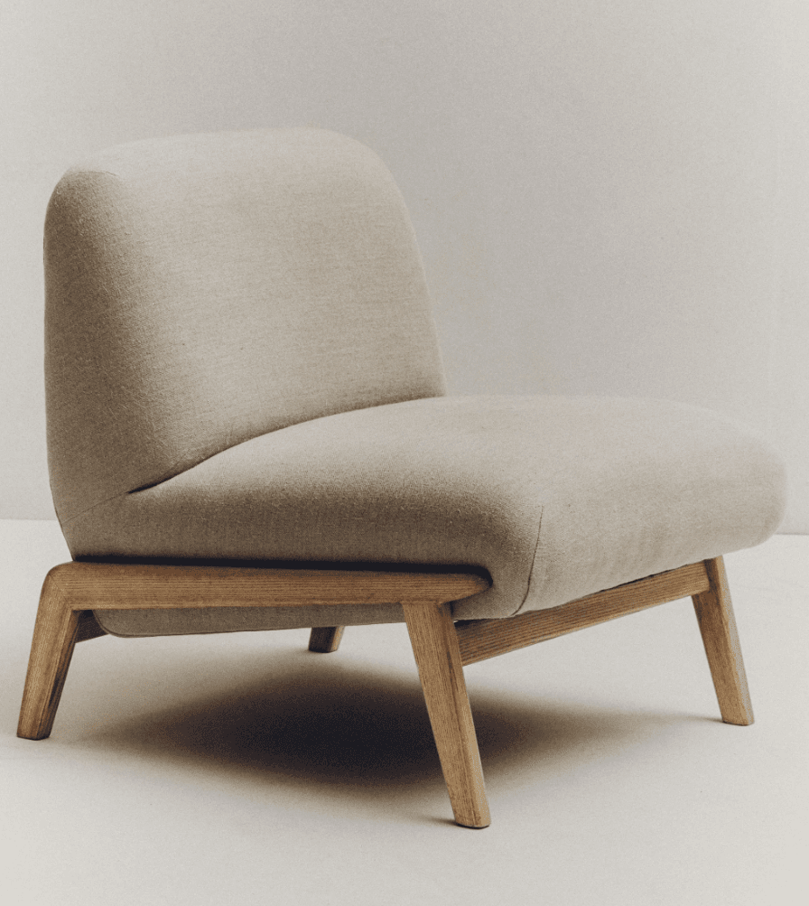 A low-profile armchair with a rounded, upholstered seat and backrest, supported by a minimalist wooden frame. The armchair features a fabric upholstery that appears to be a light beige or off-white color and a visible wooden base with four angled legs. — 3D model