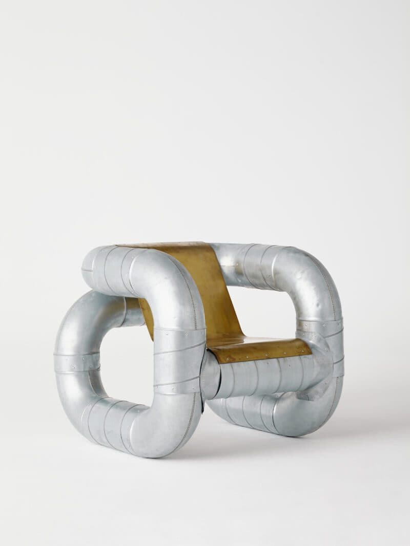 An avant-garde chair constructed from large diameter, curved metal pipes joined together to form a unique, sculptural base and frame. The seating and backrest elements appear to be made from a flat sheet of metallic material, possibly brass or a similar alloy, integrated within the pipe structure. The metal pipes exhibit visible seams and rivets, suggesting a construction method similar to ductwork or industrial piping. The overall aesthetic is industrial and sculptural. — 3D model