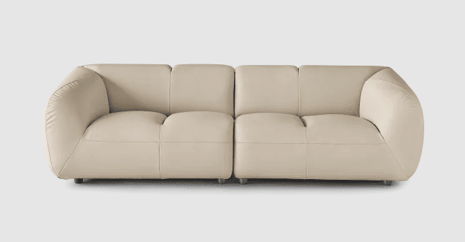 A contemporary four-seat sofa with a low profile and rounded arms. The sofa is upholstered in a light beige or cream-colored material, likely leather or a high-quality faux leather, and features prominent stitching that delineates the seat and back cushions. The design is modular, with two distinct seating sections joined together. It is supported by small, dark, cylindrical feet. — 3D model