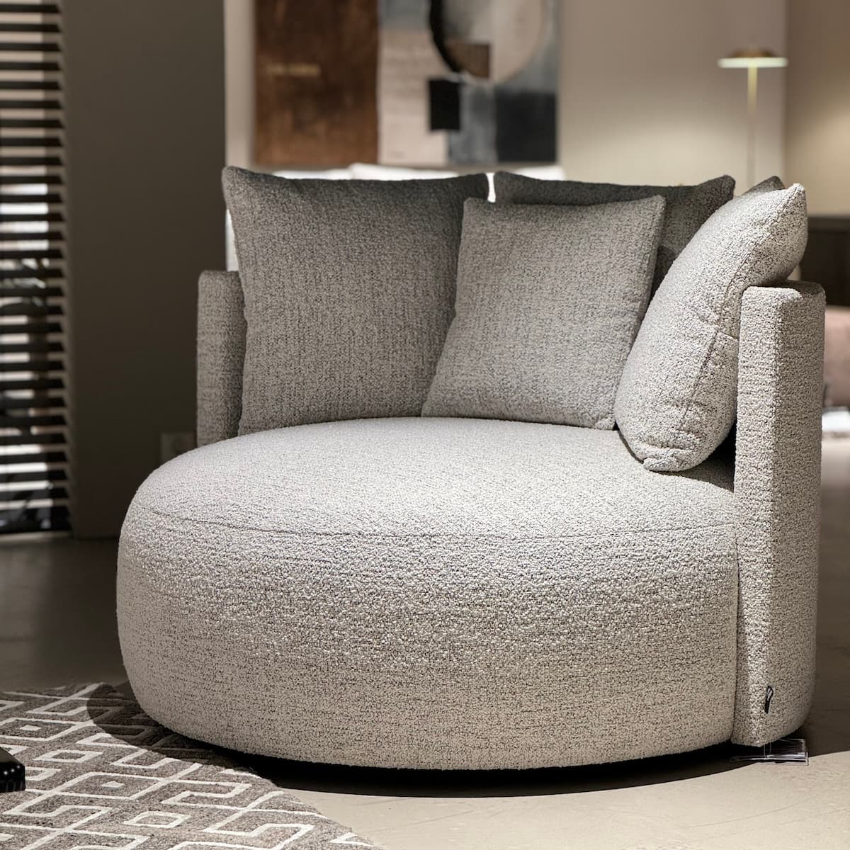 A contemporary armchair with a rounded, enveloping silhouette. It features a textured boucle fabric upholstery in a light grey hue, deep seat, and multiple plush back cushions, one of which is angled. The design suggests comfort and a modern aesthetic. — 3D model