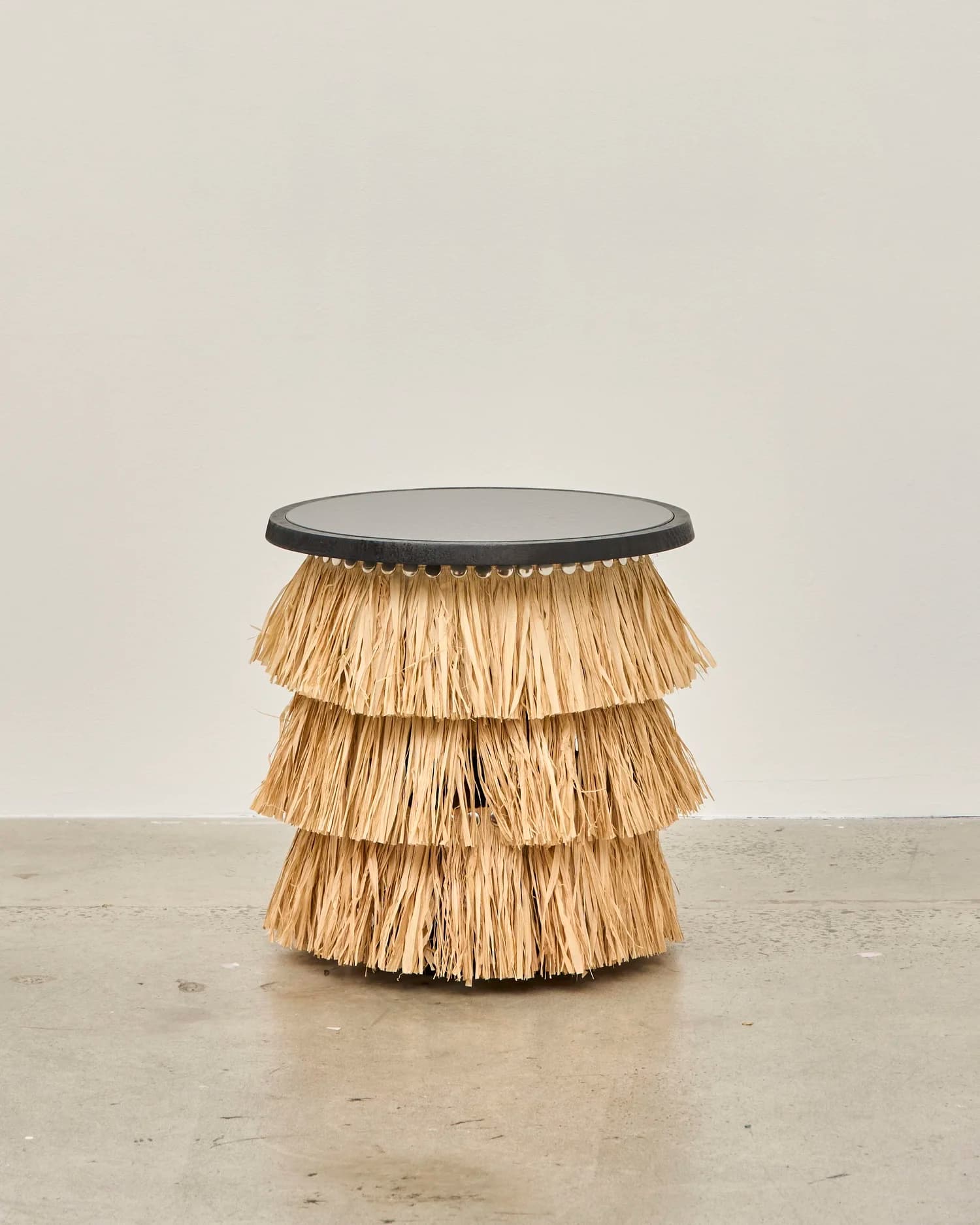 A compact side table featuring a dark, round tabletop supported by a tiered base made of natural, straw-like fibers. — 3D model