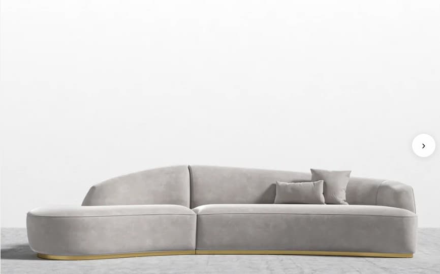 A contemporary curved modular sofa upholstered in light gray velvet, featuring an integrated backrest and armrests, supported by a recessed polished brass plinth base. It includes two matching throw pillows. — 3D model