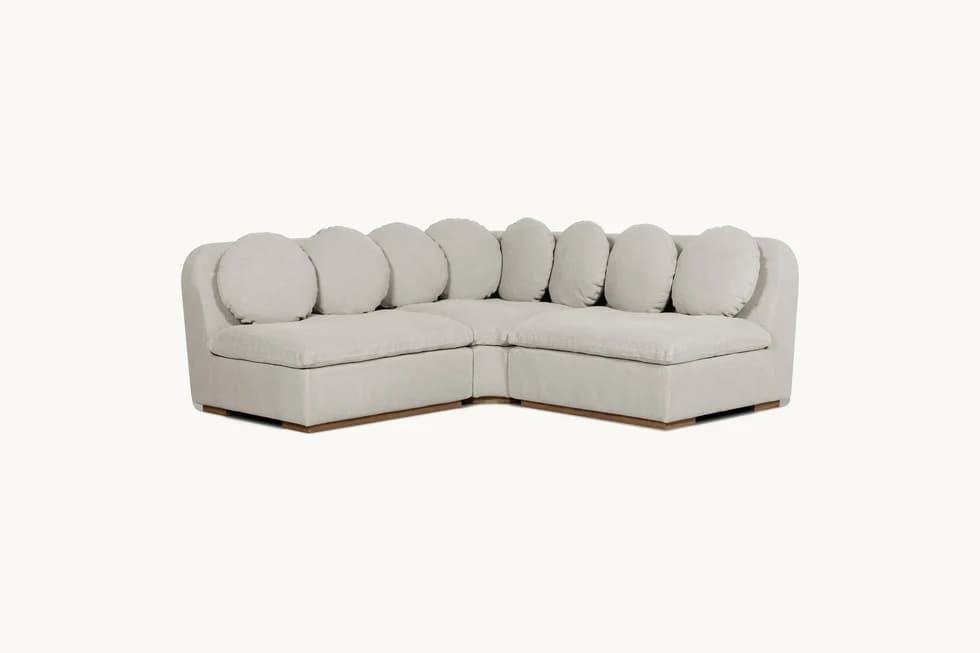 A modular sectional sofa with a low profile and rounded arms. It features a light-colored fabric upholstery and is accented by multiple large, round throw pillows along the back. The base of the sofa appears to be a dark wood. — 3D model