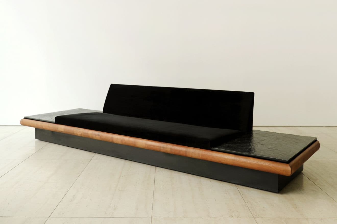 A long, low, contemporary sofa featuring a black velvet upholstered seat and a gently curved backrest, flanked by integrated dark, textured stone side tables, all set within a dark rectangular base with a continuous rounded wooden trim. — 3D model
