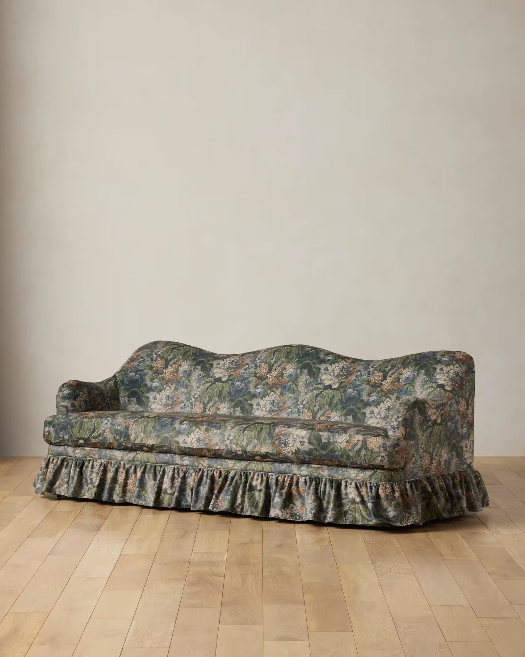A three-seater sofa featuring a distinctive scalloped backrest and a fully upholstered frame. It is covered in a dark floral fabric with prominent green leaves, blue and white flowers, and hints of orange. The sofa has rolled arms and a gathered, ruffled skirt concealing its legs. — 3D model