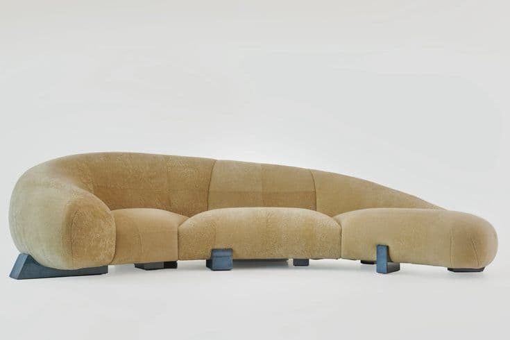 A large, curved sectional sofa with a plush, textured upholstery in a light brown or beige hue. The sofa features rounded armrests and backrests, with a modular design creating distinct seating sections. It is supported by several blocky, dark blue or gray legs with a geometric, angular profile. — 3D model