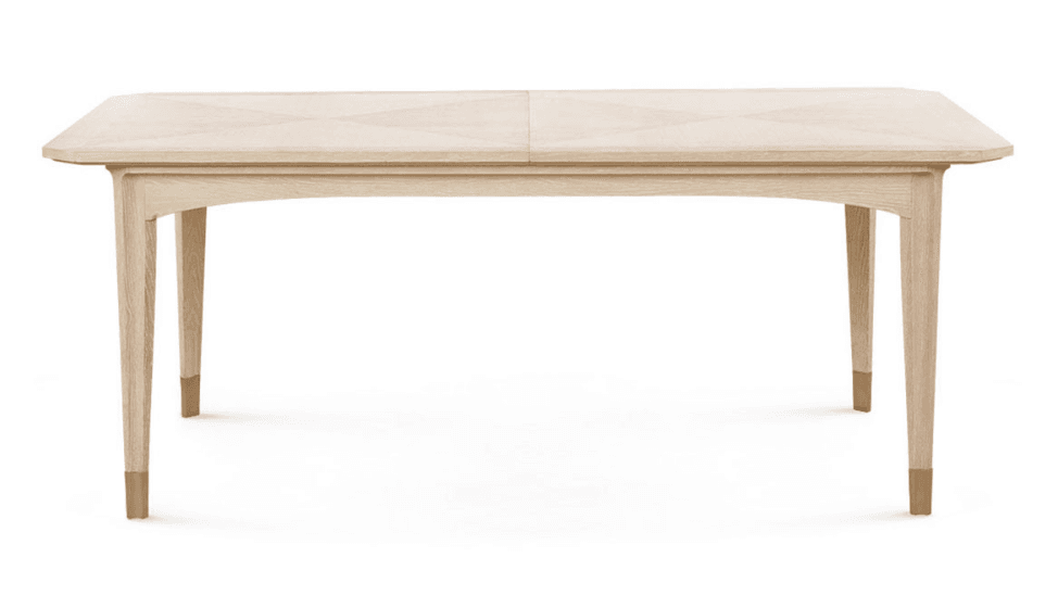A contemporary rectangular dining table made of light-toned wood, featuring a multi-panel top with a subtle geometric veneer pattern and a central seam indicating an extendable design. It stands on four tapered legs, each finished with a polished brass cap. — 3D model