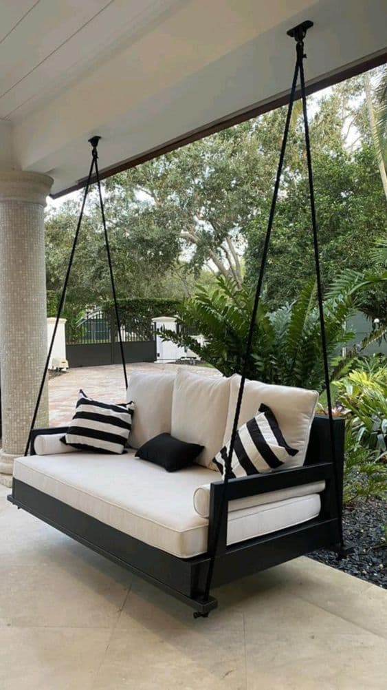A modern suspended outdoor daybed featuring a rectangular black wooden frame, thick white upholstered cushions, and several throw pillows. The daybed is hung by four black metal rods or ropes from two overhead points, creating a swinging motion. The design is minimalist and elegant, suitable for a contemporary outdoor living space. — 3D model