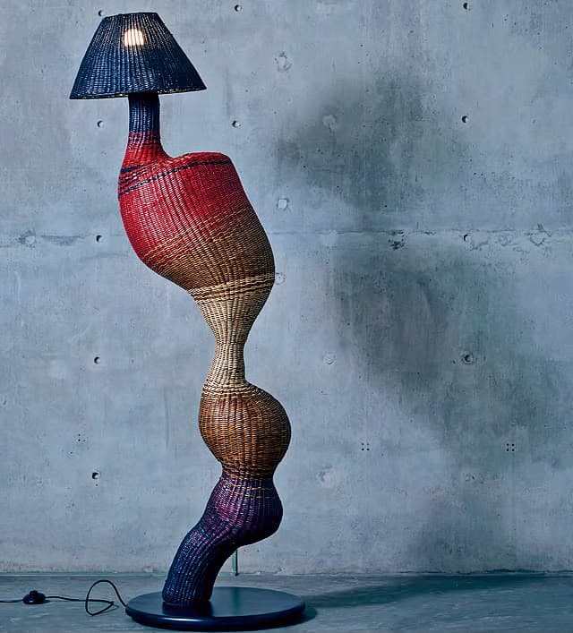 A tall, sculptural floor lamp featuring an organic, serpentine body constructed from multi-colored woven rattan or wicker. The body transitions through shades of red, brown, beige, and dark blue/purple. It is topped with a black conical mesh shade and stands on a flat, circular dark base. — 3D model
