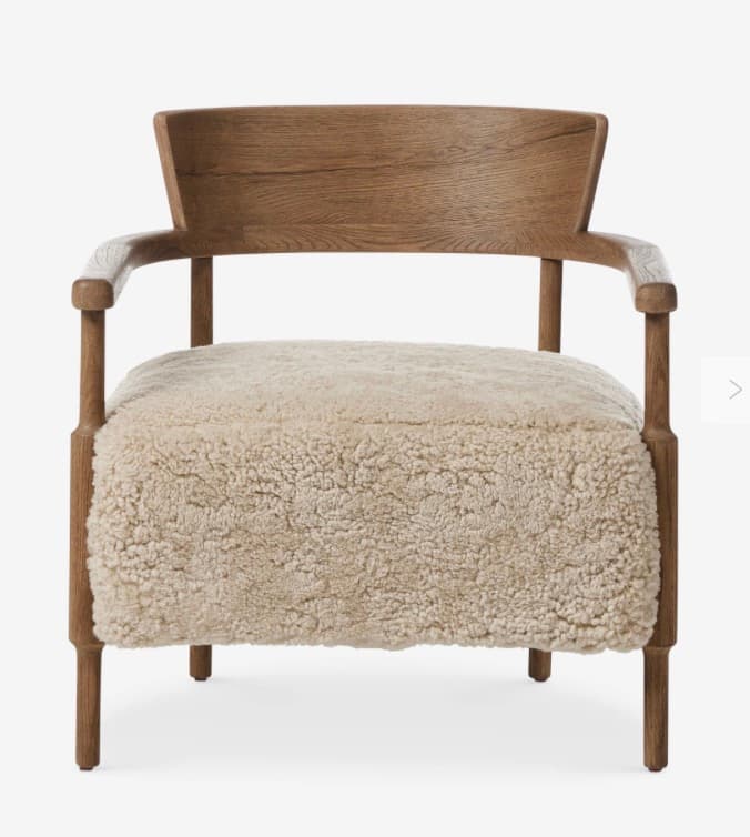 A contemporary armchair featuring a light brown wooden frame and a plush, light beige boucle or sherpa upholstered seat and backrest. The frame includes integrated armrests and subtly shaped legs. The backrest is a curved wooden panel. — 3D model