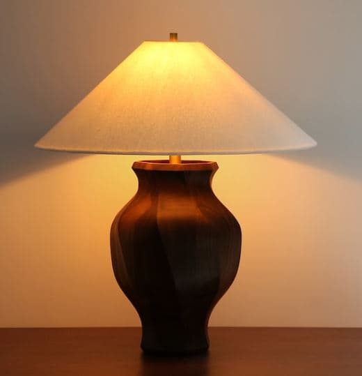 A table lamp featuring a dark, vase-shaped wooden base and a light-colored conical fabric shade. The lamp emits a warm, diffused light and is suitable for ambient illumination. — 3D model