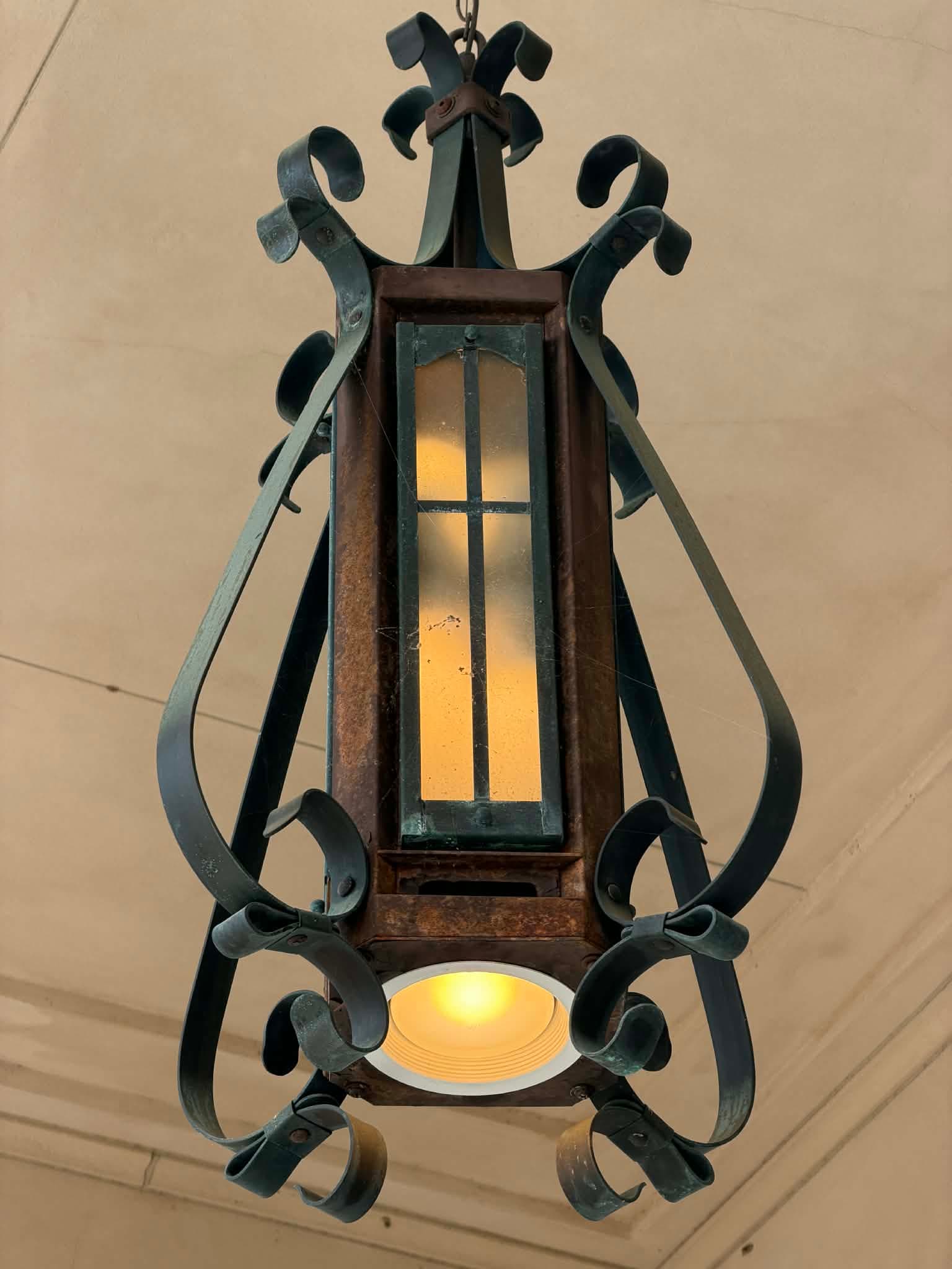 An ornate hanging lantern made of dark wrought iron and rusted metal, featuring frosted glass panels and intricate scrollwork. It has a downward-facing light source and emits light through the panels. — 3D model
