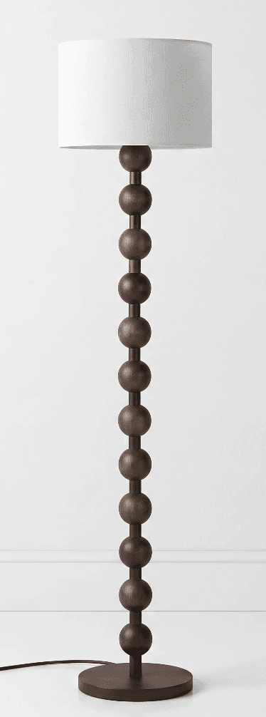 A tall floor lamp featuring a distinctive dark brown wooden stem composed of a series of stacked, spherical beads. It rests on a flat, round wooden base and is topped with a simple white cylindrical fabric drum shade. — 3D model