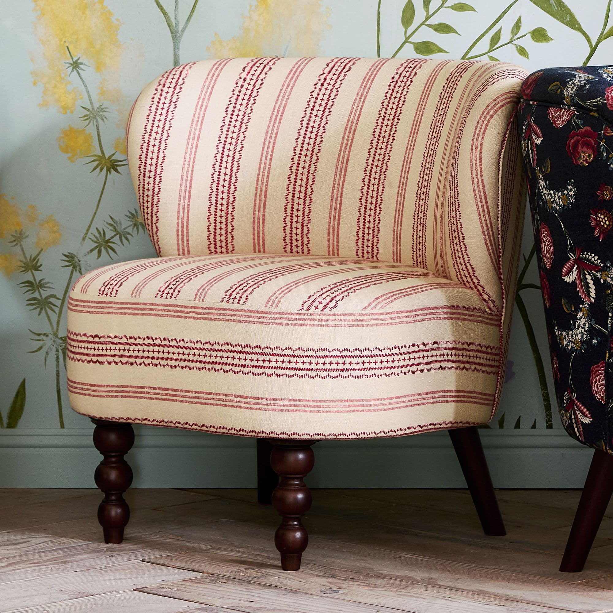 A fully upholstered accent chair with a curved back and seat. The chair features a cream-colored fabric with a repeating pattern of thin red stripes and a more intricate red geometric pattern interspersed with the stripes. It is supported by four legs, with the front two legs having a turned, bulbous design and the rear legs being tapered and darker in color. The overall design suggests a vintage or traditional aesthetic. — 3D model