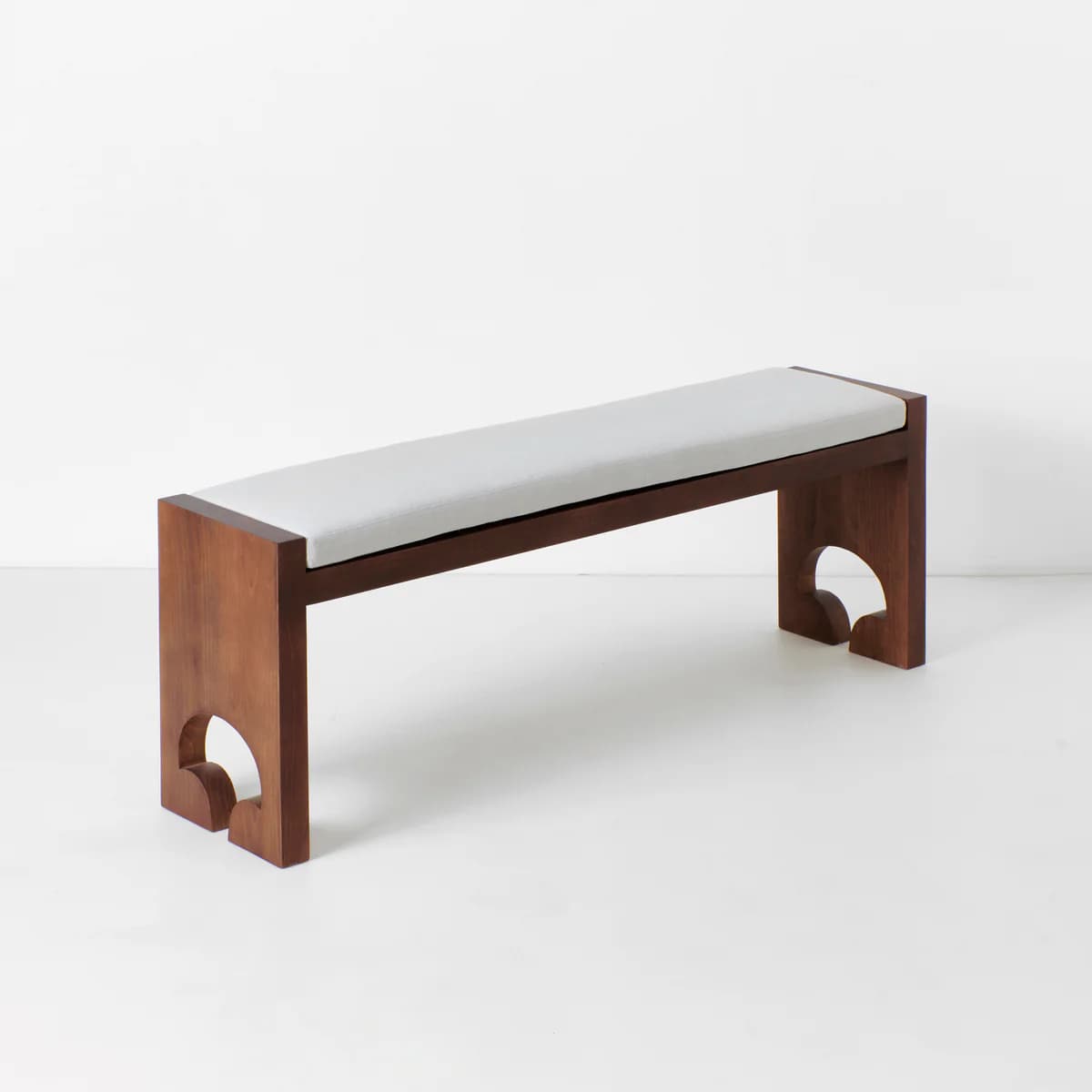 A wooden bench with a upholstered seat. The bench has a minimalist design with straight lines for the frame and a rectangular, cushioned top. The legs are thick and feature a distinctive arched cutout at the base, creating a decorative element. The overall aesthetic suggests a modern or contemporary style. — 3D model