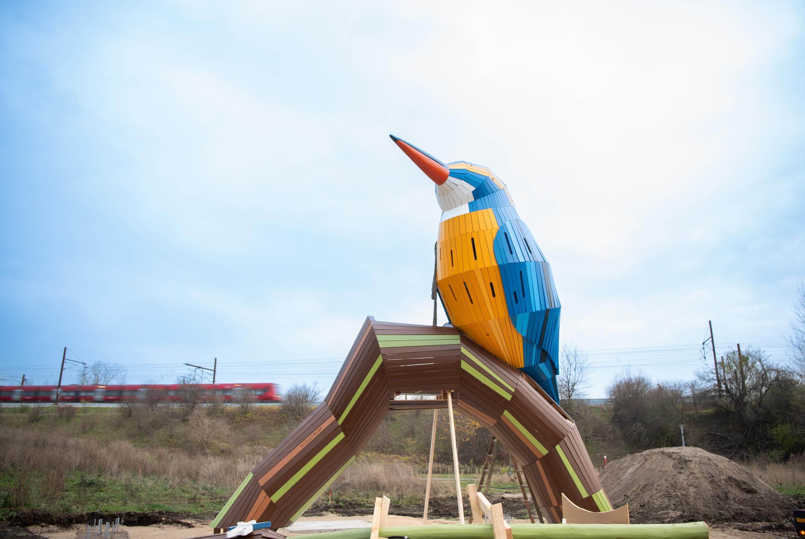 A decorative sculpture depicting a kingfisher bird perched on a branch, crafted from multiple segmented pieces of wood. The bird features a vibrant blue, orange, and white palette, while the branch is predominantly brown with green and lighter brown accents, all finished with a smooth, glossy appearance. — 3D model