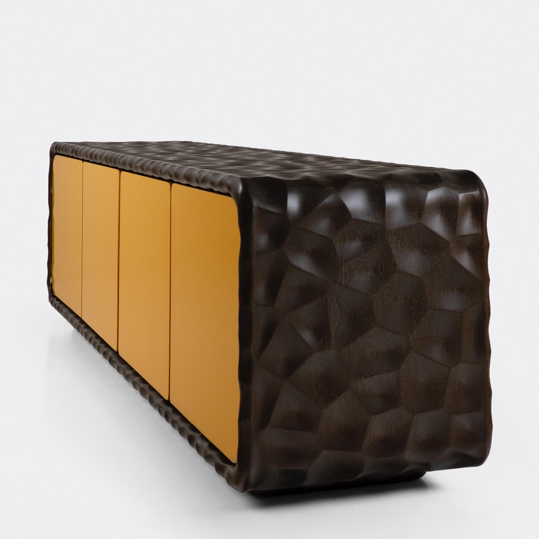 A long, low-profile contemporary sideboard featuring a deeply textured, dark brown wooden exterior that wraps around the top, sides, and bottom. The front of the sideboard is composed of four flush, smooth, ochre-colored lacquered doors, creating a striking contrast with the organic, sculpted wood frame. The texture on the wood resembles a faceted, geometric pattern or carved rock, giving the piece a sculptural and avant-garde aesthetic. The piece appears to rest directly on the floor without visible legs. — 3D model