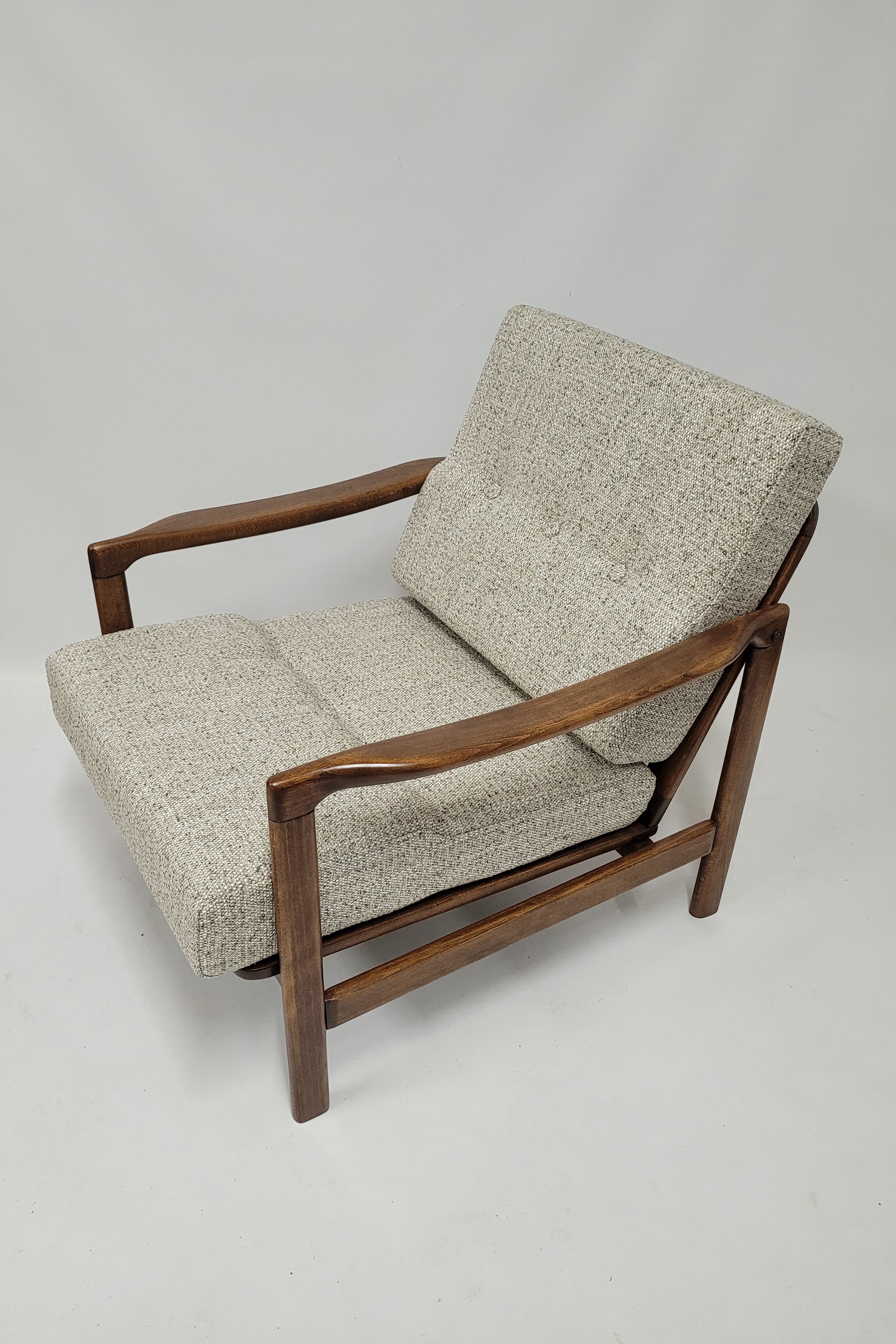 A mid-century modern armchair with a wooden frame and upholstered seating and backrest. The frame appears to be made of dark wood, possibly walnut or teak, with a smooth finish. The arms are gently curved, flowing from the back legs to the front. The seat and back cushions are upholstered in a textured fabric with a speckled pattern in light gray and off-white tones. The back cushion is tufted with visible buttons. The construction features clean lines and a minimalist aesthetic. — 3D model