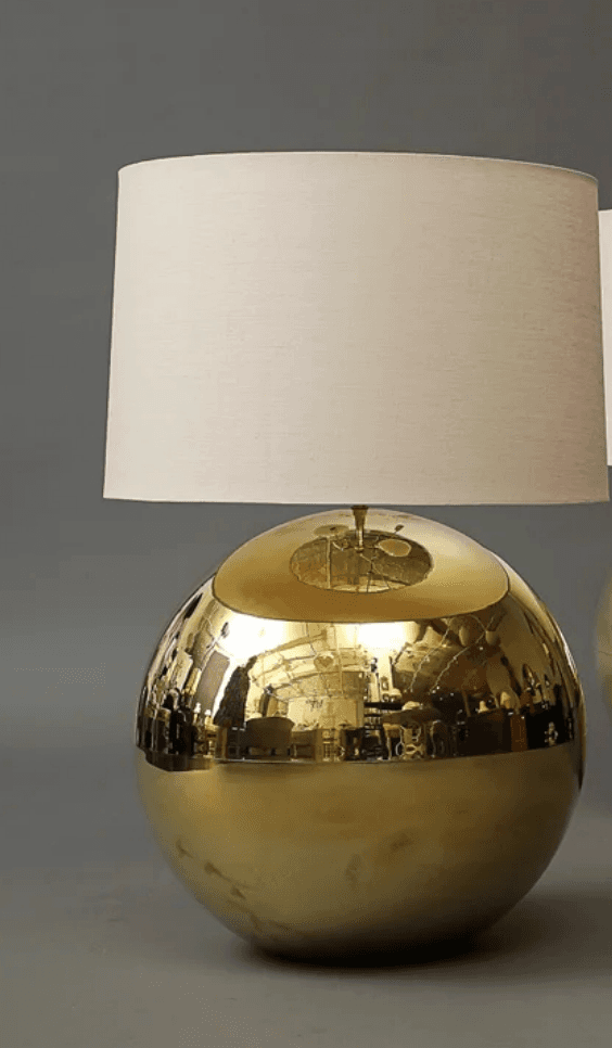 A table lamp featuring a spherical, highly polished brass-toned metal base and a plain, cylindrical off-white fabric lampshade. The base exhibits strong reflections of its surroundings, indicating a lustrous, mirror-like finish. The design is minimalist, focusing on the material and geometric form. — 3D model