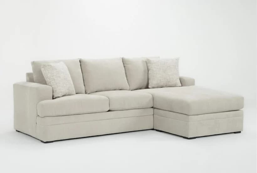 A contemporary L-shaped sectional sofa featuring a right-hand facing chaise. It is upholstered in a light beige or off-white fabric, presenting a clean and neutral aesthetic. The sofa includes loose seat cushions, three loose back cushions, and two smaller toss pillows with a subtle textured pattern. The armrests are broad and squared, contributing to its modern, relaxed design. The base of the sofa is low-profile with small, dark, recessed feet. — 3D model