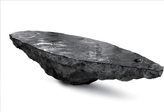 A unique coffee table designed to resemble a floating rock formation with a dark, textured surface, possibly slate or a similar stone, topped with a reflective, water-like surface exhibiting ripple patterns. The underside of the rock shows rough, unworked geological features. The overall aesthetic suggests a naturalistic and artistic design. — 3D model