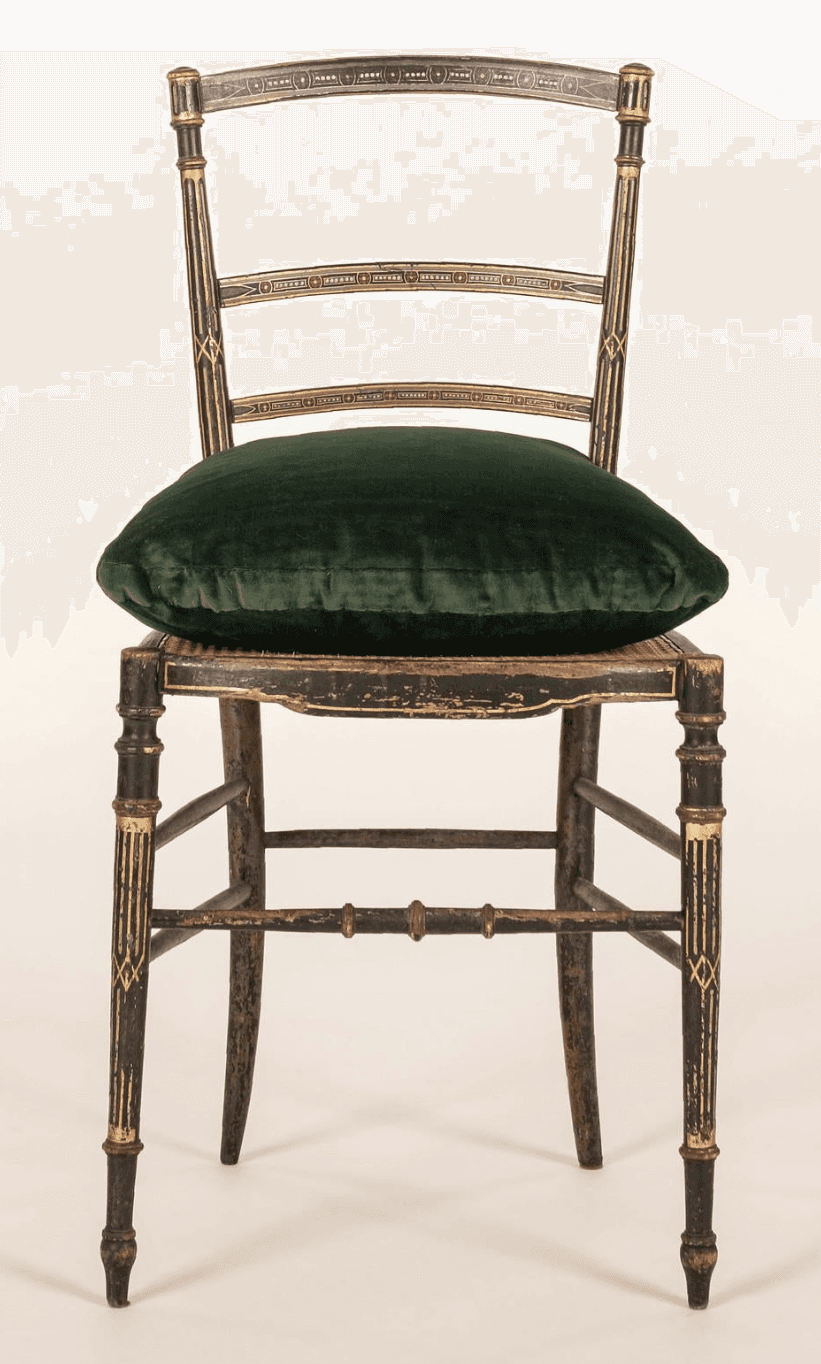 An antique Regency style side chair featuring an ebonized (black lacquered) wood frame with extensive gilt decoration. The chair has a slat back design with three horizontal slats adorned with geometric gilt patterns, complemented by reeded and gilt uprights and finials. The seat appears to be caned, currently topped with a loose, plush dark green velvet cushion. The front legs are turned with gilt accents, while the splayed rear legs are plain. Stretchers connect the legs, some with ring-turned details, and the ebonized finish shows visible distressing and wear. — 3D model