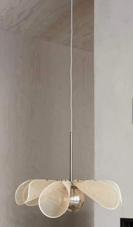 A contemporary pendant light featuring a shade composed of multiple off-white, petal-shaped fabric elements. These elements are arranged radially around a central brass stem, connected by small brass studs, creating an organic, floral silhouette. A single exposed light bulb is positioned at the center, underneath the fabric petals. The fixture hangs from a single white electrical cord connected to the brass stem. — 3D model