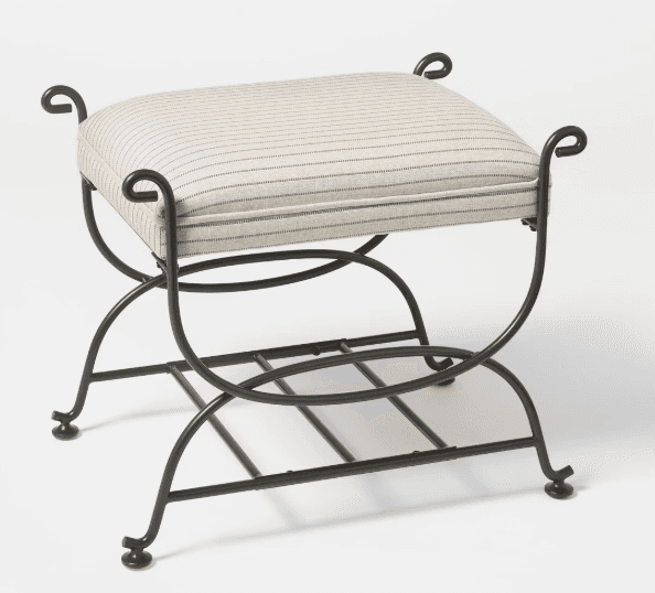A rectangular upholstered stool featuring a dark-finished wrought iron frame with an X-shaped base. The seat cushion is upholstered in a light-colored fabric with thin dark pinstripes. The frame incorporates decorative scrolled finials at each corner of the seat and includes a slatted metal shelf near the base, suggesting additional utility. The design combines traditional elements with a functional form. — 3D model