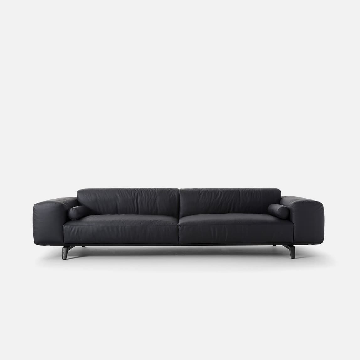 A contemporary three-seater sofa upholstered in black leather, featuring a low profile, clean lines, and broad, low arms. It includes two integrated cylindrical bolster pillows, one at each end. The sofa rests on slender, dark-finished metal legs, contributing to its modern minimalist aesthetic. — 3D model