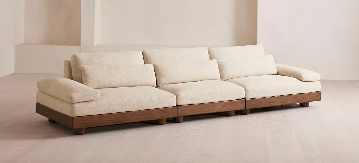 A sectional sofa with three distinct seating modules. Each module features plush, upholstered cushions for the seat and back, and a single armrest on the outer side of the leftmost and rightmost modules. The base of the sofa is a continuous wooden frame with a rich, dark wood finish, supported by short, cylindrical legs. — 3D model