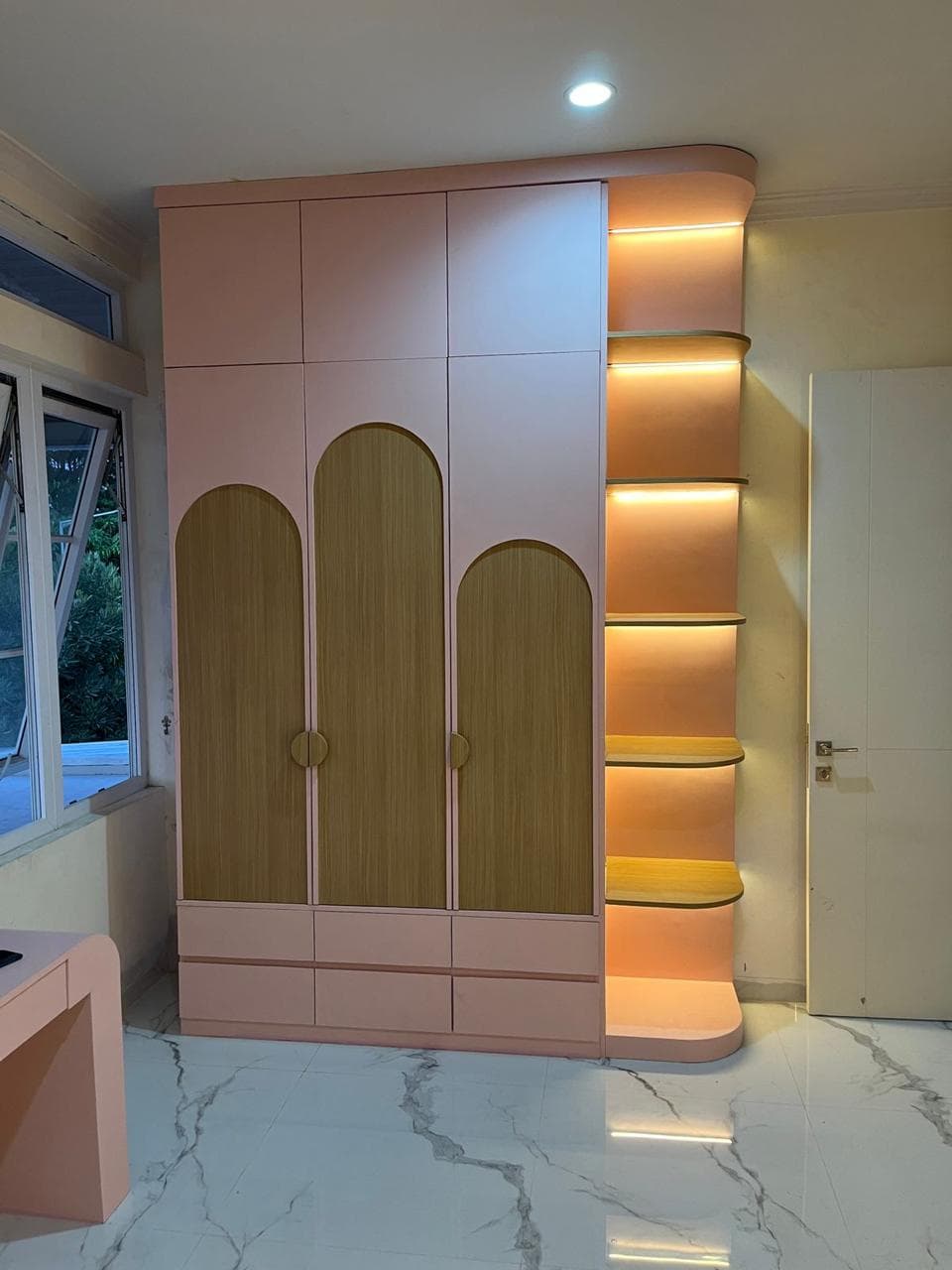 A modern, tall storage unit in pink with natural wood accents, featuring multiple closed cabinet doors, base drawers, and integrated, illuminated corner shelves on one side. — 3D model