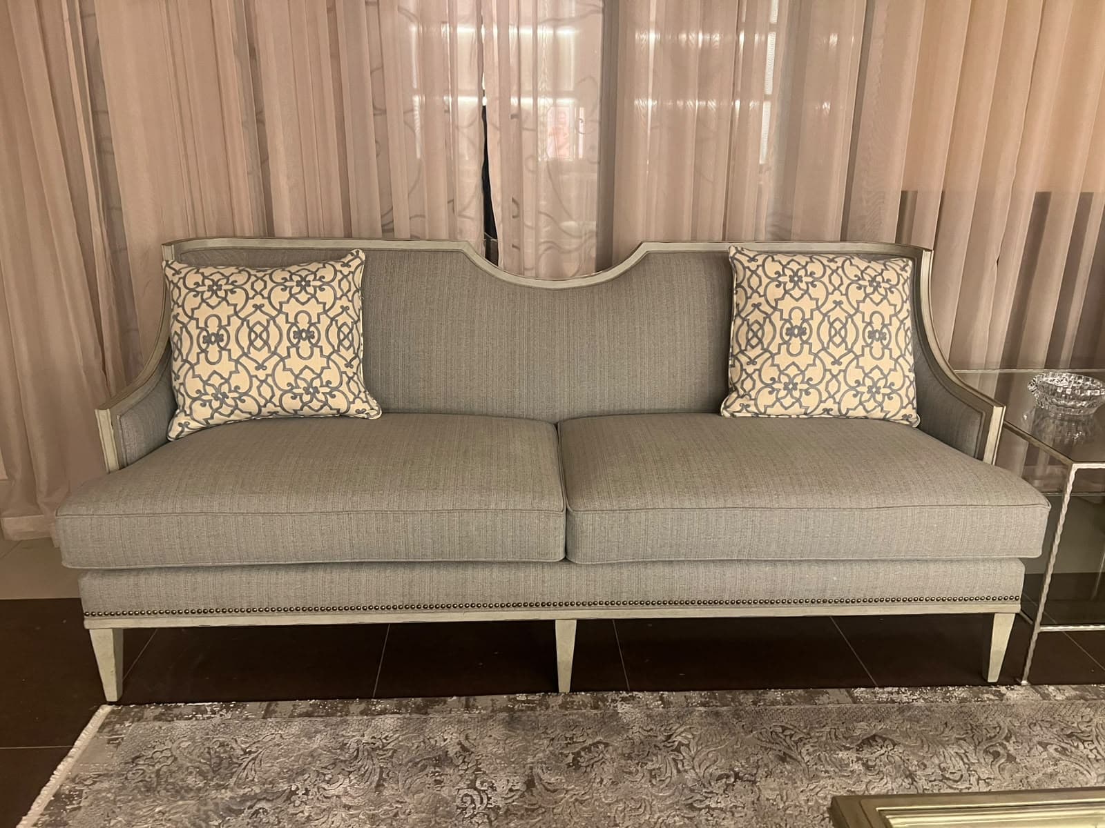 A light grey upholstered sofa featuring an exposed light-toned wooden frame, a gently curved backrest, and tapered legs. The sofa includes two seat cushions and two decorative throw pillows with a geometric pattern. Nailhead trim detailing is present along the base. — 3D model