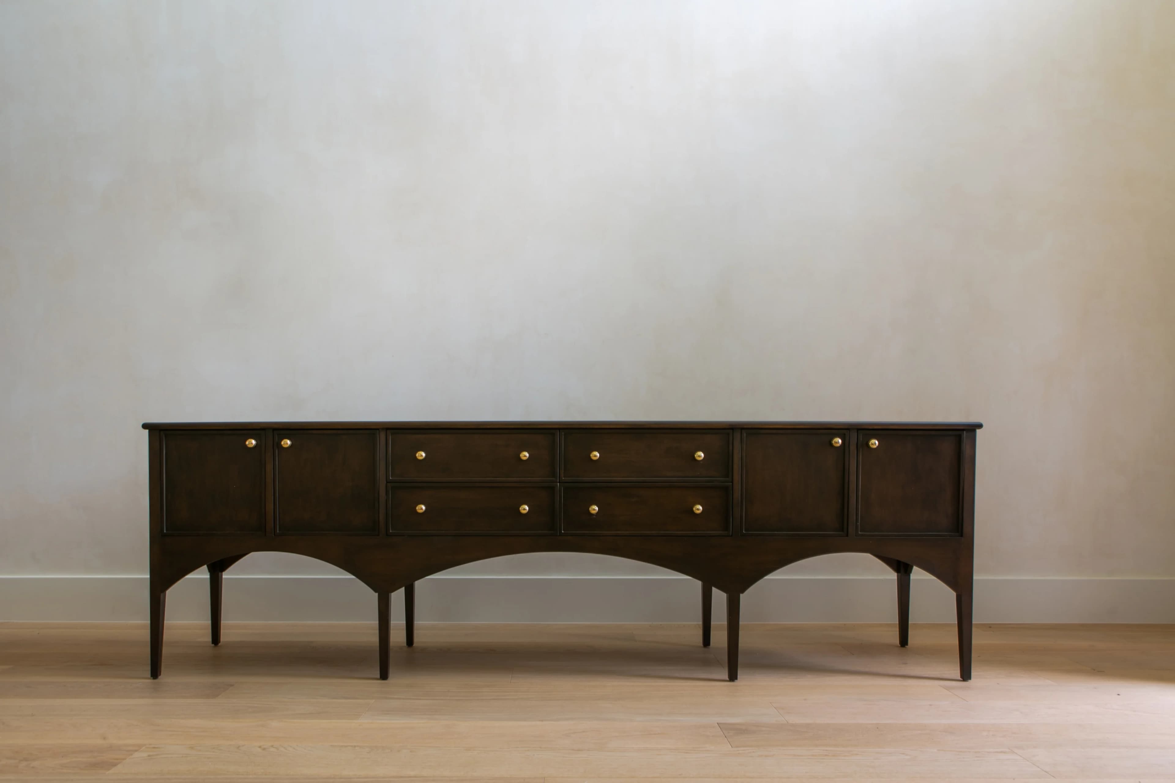 A long, low, dark-stained wooden sideboard featuring a central bank of six drawers flanked by two-door cabinets, all adorned with brass knob pulls, and supported by tapered legs with a distinctive arched apron base. — 3D model
