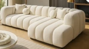 An off-white or cream-colored modular sectional sofa featuring distinctive channel tufting across all seating surfaces and backrests. The sofa is composed of multiple segments, including a long straight section and a corner chaise lounge, allowing for a versatile L-shaped configuration. Its design is characterized by soft, rounded edges and a low-profile, recessed dark wood or metal base. The sofa includes two matching boucle throw pillows, one square and one round, enhancing its plush appearance. — 3D model