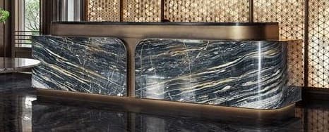 A sophisticated modern reception desk, featuring a substantial body crafted from black and gold veined marble. The desk incorporates rounded corners and two distinct front panels, accented by a metallic bronze-finished frame that wraps around the base and between the marble sections. A raised, L-shaped counter section, also in a dark, possibly stone or metallic finish, sits atop the main structure, supported by the bronze-finished frame. — 3D model