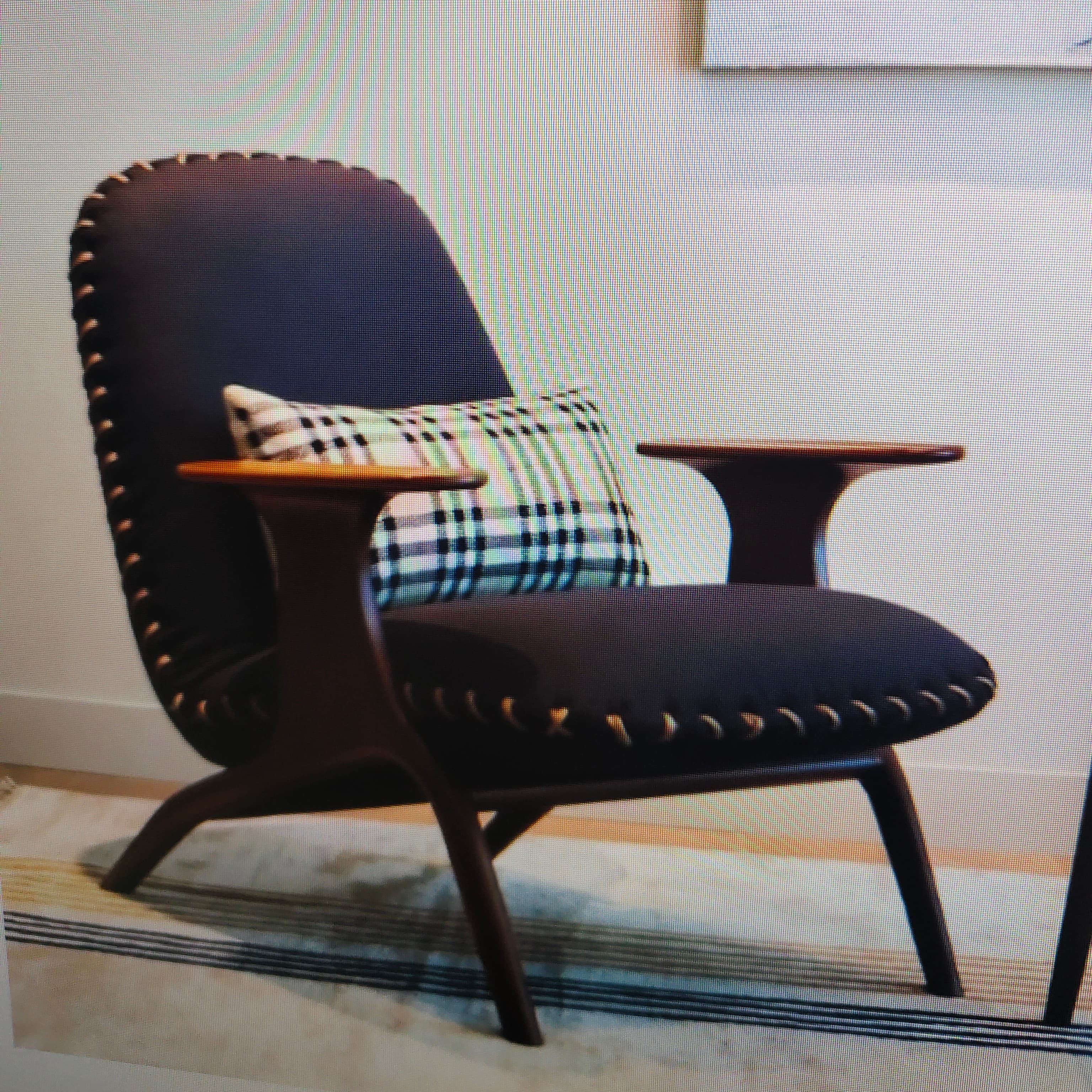 A modern lounge chair featuring a dark wooden frame with integrated, curved armrests and splayed legs. The seat and backrest are upholstered in deep navy blue fabric, accented by prominent exposed whipstitch detailing in a contrasting lighter thread along all edges. A plaid lumbar pillow rests on the seat. — 3D model