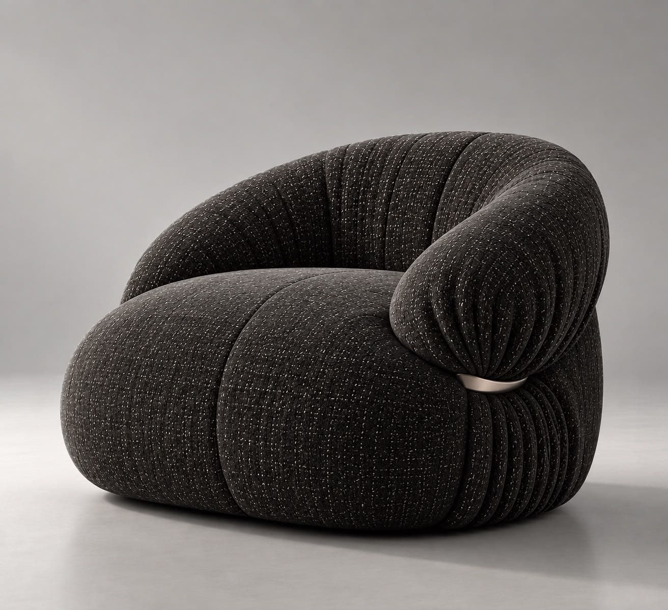A contemporary, generously padded armchair with a sculptural and organic form, upholstered in a dark charcoal grey fabric with subtle white flecks. The chair features a rounded, low-slung profile and distinct pleating or gathering of the fabric, particularly evident around the armrests and base, giving it a plush, cloud-like appearance. A single, sleek silver-toned metal ring acts as a decorative and structural detail on one side, cinching the upholstery. — 3D model