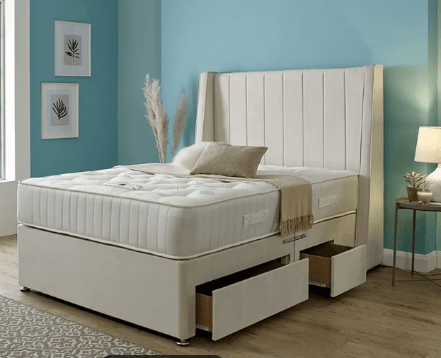 A contemporary upholstered divan bed featuring a tall, winged headboard with vertical channel tufting. The headboard and divan base are covered in a beige fabric. The divan base includes integrated storage drawers on one side and stands on short, chrome-finished feet. A mattress with visible button tufting and piped edges is placed on top of the divan base. — 3D model