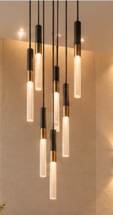 A modern multi-light pendant chandelier featuring nine individual cylindrical light sources suspended from a circular black canopy. Each light comprises a black cylindrical top, a brass band, and a frosted white or textured glass lower section that emits light. The lights hang at varying lengths, creating a dynamic, clustered arrangement. — 3D model