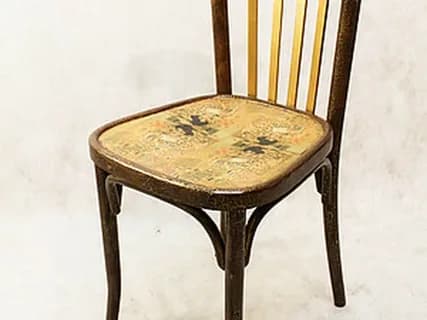 A vintage or antique wooden chair, possibly bentwood, featuring a curved backrest with vertical spindles. Some spindles are painted gold, contrasting with the dark wood frame. The seat is a solid surface with a faded, ornate pattern. The legs are curved, reflecting classic cafe or bistro chair design. — 3D model