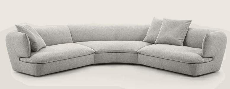 A curved sectional sofa upholstered in a light grey textured fabric. It features a low profile with rounded armrests and deep seating. Two matching throw pillows are placed on the sofa. — 3D model