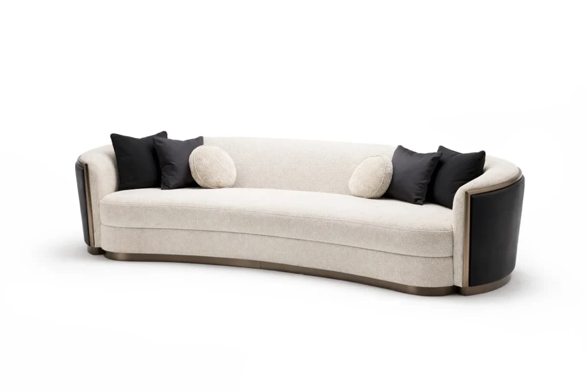 A contemporary curved sofa featuring a cream-colored boucle fabric upholstery. The sofa has a distinct curved silhouette and integrated armrests that are partially clad in dark, possibly black, leatherette or faux leather, framed by a brass or bronze-finished metal trim. A continuous metal trim also accents the base of the sofa. It comes with two black square throw pillows and two circular, cream-colored boucle pillows. — 3D model