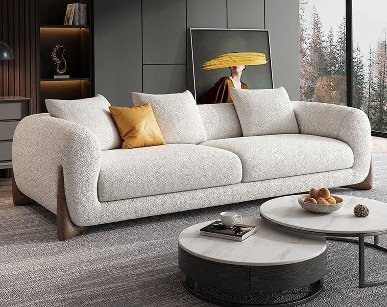 A modern, three-cushion sofa with a curved armrest and a textured fabric upholstery. It features thick, plush seat and back cushions, and two decorative throw pillows. The sofa is supported by dark, angular legs that contrast with the lighter upholstery. A small bowl of decorative fruit is placed near the front right leg. — 3D model