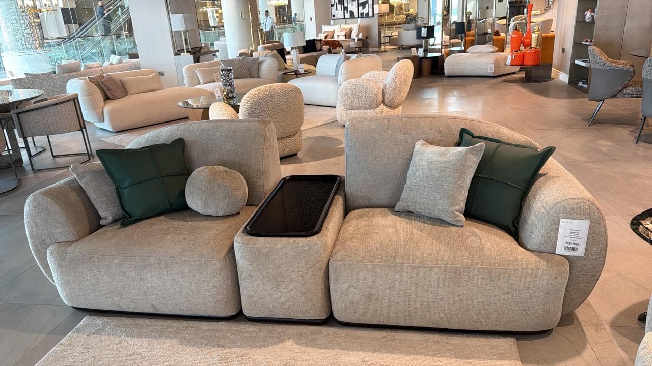 A modular sectional sofa with a low profile and plush upholstery. The sofa features multiple seating units, including a corner piece and armless sections, arranged in a linear configuration. It is adorned with several throw pillows in varying shades of green and grey, and a black tray is integrated into the armrest or between seating sections. The overall design is contemporary and appears to be upholstered in a textured fabric. — 3D model
