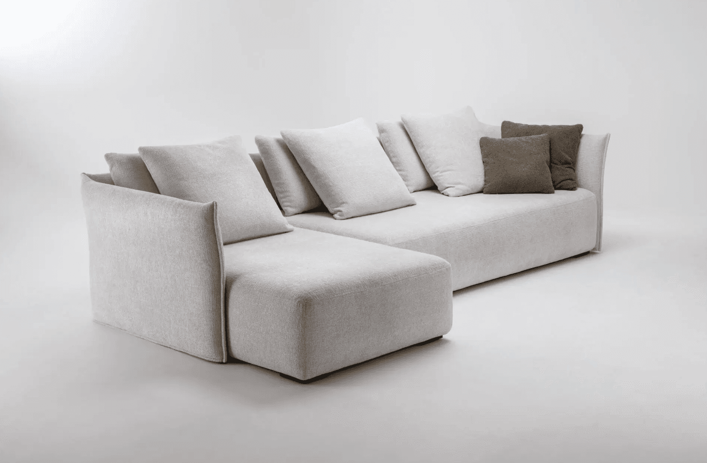 A sectional sofa with a chaise lounge extension. The sofa features deep seating and is upholstered in a textured fabric. It is adorned with multiple throw pillows, some in a lighter shade matching the sofa and others in a darker, contrasting brown. The design appears modern with clean lines and soft, rounded edges on the arms. — 3D model